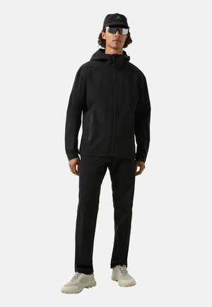 Veste noire pour homme avec capuche, poche frontale zippée, associée à un pantalon noir et des chaussures gris clair. Les accessoires comprennent une casquette noire et des lunettes de soleil.