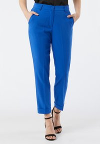 Pantalon bleu sur mesure en tissu lisse, doté d'une coupe droite, de poches latérales et d'ourlets retroussés. Porté avec des talons noirs à lanières.