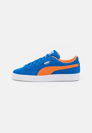 Zapatilla Puma de ante azul con franja naranja y forro interior, suela blanca y cordones, vista de lado sobre un fondo blanco.