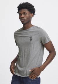 Grå kortærmet t-shirt i et blødt materiale med rund hals, med en lille sort grafisk detalje på brystet. Bæres med blå jeans.