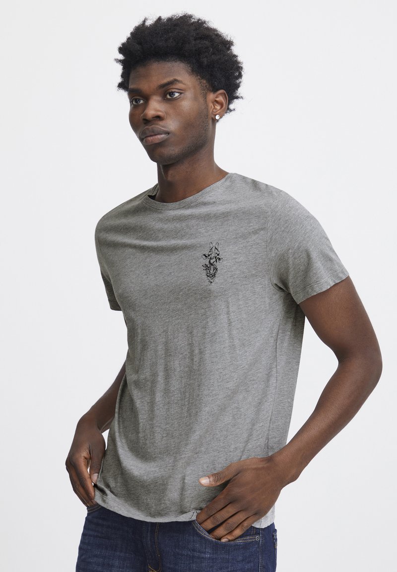 Grå kortærmet t-shirt i et blødt materiale med rund hals, med en lille sort grafisk detalje på brystet. Bæres med blå jeans.