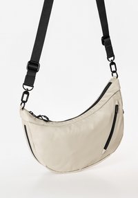 Beige Schultertasche mit geschwungenem Design, aus weichem Stoff gefertigt. Verfügt über eine schwarze Reißverschlusstasche und einen verstellbaren schwarzen Riemen.