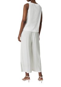 Top sans manches en lin blanc associé à un pantalon large en lin clair, avec des fentes latérales. Le mannequin porte des sandales blanches à talons.