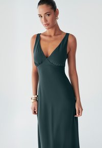 Varrukateta sügava V-kaelusega kleit tumerohelisest kangast. Kehaosa on istuv ja läheb laienevaks, voolava seelikuni. Sile tekstuur, minimalistlik disain, peened õmbluse detailid.