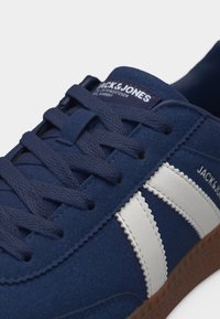 Σκουρό μπλε αθλητικό παπούτσι με λευκές πλευρικές ρίγες, υφασμένο καφέ πέλμα και σκουρό μπλε κορδόνια, με επωνυμία "JACK & JONES" στο πλάι και στη γλώσσα.