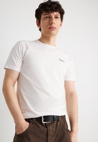 Νεαρός άνδρας που φοράει λευκό μπλουζάκι με στρογγυλή λαιμόκοψη και μικρό μαύρο λογότυπο, καφέ παντελόνι και μαύρη ζώνη, στέκεται με τα χέρια στις τσέπες δίπλα σε λευκό τοίχο.