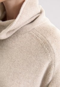 Beżowy sweter dziany z dekoltem typu cowl, charakteryzujący się gładką teksturą oraz subtelnymi detalami w postaci przeszyć wzdłuż ramion i rękawów.