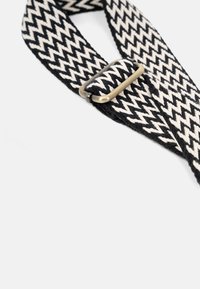 Gewebtes Band in Schwarz und Creme mit einem Zickzackmuster. Verfügt über eine Messing-Schnalle zur Anpassung. Flaches Design mit strukturiertem Oberflächen.