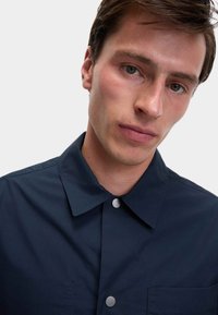 Mørkeblå skjorte med knapper lavet af glat stof, med krave, sølvfarvede knapdetaljer og en brystlomme på venstre side.