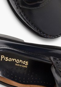 Zapato de cuero negro con un acabado suave, que presenta costuras visibles y plantillas perforadas, etiquetado "Pisamonas Hecho en España."