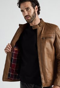 Mann in brauner Lederjacke mit kariertem Innenfutter, hält sie offen, um das Innere zu zeigen, gekleidet in einem schwarzen Hemd und dunklen Jeans.