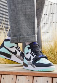 Černé a mátově zelené vysoké tenisky s bílými akcenty, texturovaná šedá mezipodešev a modré Nike logo, doplněné šedými kostkovanými kalhotami.