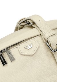Una borsa a mano in pelle color crema con superficie testurizzata, hardware argentato, chiusura con zip e un accento con logo. Inclusa una tracolla regolabile.