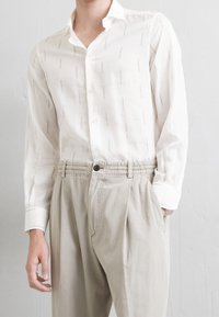 Mężczyzna w białej koszuli zapinanej na guziki z subtelnym wzorem, włożonej w beżowe, plisowane spodnie, stojący na jasnym tle.