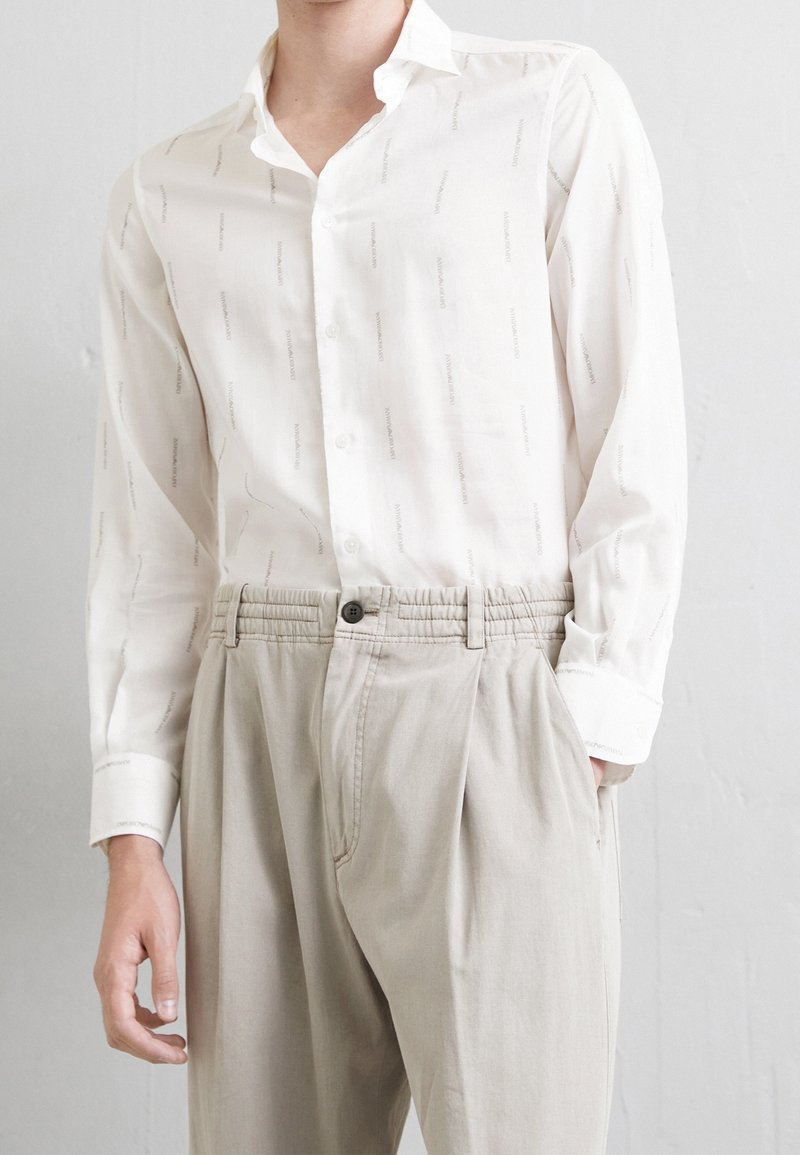 Mężczyzna w białej koszuli zapinanej na guziki z subtelnym wzorem, włożonej w beżowe, plisowane spodnie, stojący na jasnym tle.