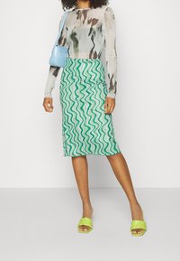 Průsvitný dlouhý rukáv vrchní díl s abstraktními vzory v šedé a hnědé, v kombinaci s zelenou tužkovou sukní s vlnitým vzorem a limetkově zelenými sandály na podpatku.