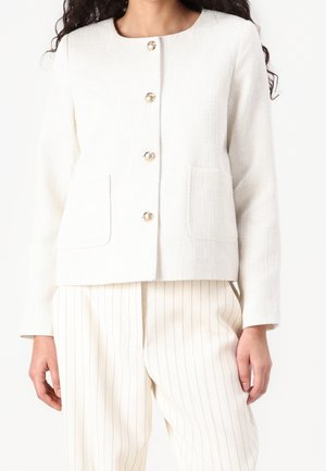 Femme portant une veste blanche texturée avec quatre boutons dorés et des poches plaquées, associée à un pantalon crème à fines rayures.