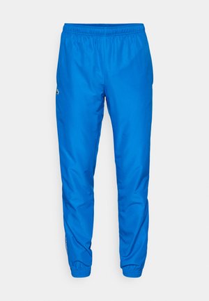 Pantalons de sport bleus avec une taille élastique, jambes fuselées et texture lisse. Présente un logo sur le côté et des rayures blanches discrètes.