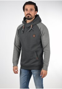 Sudadera gris oscuro con mangas gris claro, con un bolsillo tipo canguro en el frente, capucha con cuerda ajustable y un emblema de cuero.