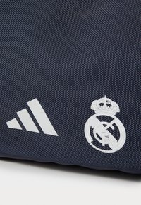 Mørkeblå stoftaske med hvid Adidas-logo og Real Madrid-emblem, præsenteret med en struktureret overflade og minimalistisk design.