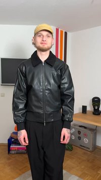 Juoda dirbtinės odos bombonešio striukė su kailio apykakle, megztais rankogaliais ir apačia; modelis vilki juodus kelnes, minimalistinė aplinka.