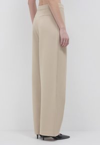 Pantaloni ampi beige con tasche posteriori a fessura, indossati con décolleté neri a punta e tacco kitten, mostrati di lato e posteriormente su uno sfondo neutro.
