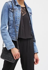 Veste en denim avec deux poches poitrine, couleur bleu clair, portée sur une combinaison noire avec des détails en dentelle. Sac bandoulière noir avec des clous.