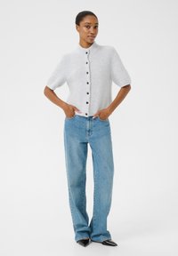 Helehallist lõngakootud lühikeste varrukatega kardigan kõrge kaelusega ja mustade nööpidega, koos siniste sirgete teksade ja terava musta kingadega.