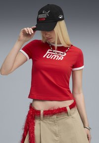 Röd tajt t-shirt med vit kant som har en djärv "PUMA"-logotyp. Svart basebollkeps med logobeklädnad. Laptop beige byxor med ett fluffigt rött bälte.