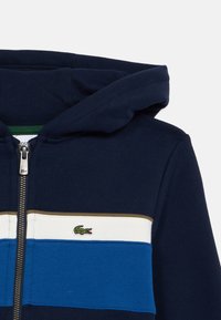 Hanorac navy cu fermoar, cu dungi orizontale albe și albastre și un mic logo verde de crocodil pe piept.