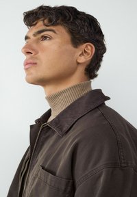 Brązowa kurtka kordowa z wywijanym kołnierzem, noszona na beżowym prążkowanym golfie. Włosy są ciemne i kręcone, lekko skierowane ku górze.