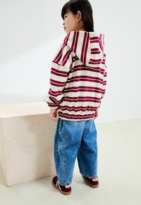 Veste à capuche rayée en crème et bordeaux, associée à un jean bleu ample et des chaussures bordeaux avec des accents roses. Tissu doux, coupe décontractée.