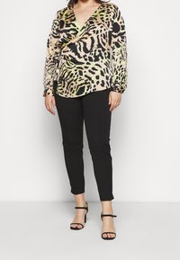 Løveprikket bluse i beige, grøn og sort med omslagdesign og lange ærmer. Sammen med tætsiddende sorte bukser og strophæle.