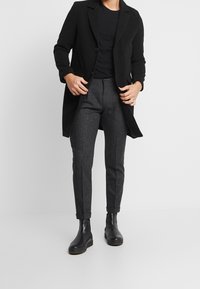Zwarte mantel met geknipte revers, knoopsluiting en voorzakken, gecombineerd met donkergrijze broek en zwarte leren enkellaarzen.