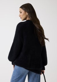 Pull noir texturé avec une coupe ample, des manches larges et un dos légèrement allongé, associé à un jean bleu.