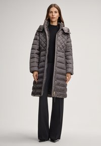 Cappotto grigio imbottito con design trapuntato, colletto grande, dettagli in metallo dorato e zip laterale. Indossato sopra un top e pantaloni neri.