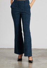 Pantalones de campana de tejido azul marino con cintura alta, bolsillos inclinados y detalles de costura visibles, combinados con tacones negros brillantes.