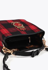 Bolso de mano a cuadros en rojo y negro con emblema bordado, correa de cuero negra, herrajes dorados y un compartimento con cremallera abierta que muestra un forro de estampado de leopardo.