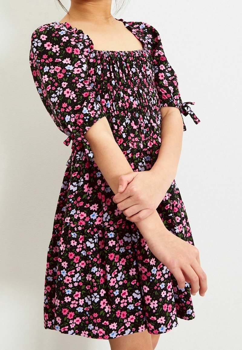 Fille portant une robe noire avec un imprimé floral rose, violet et bleu, manches bouffantes et corsage smocké, bras croisés à la taille.