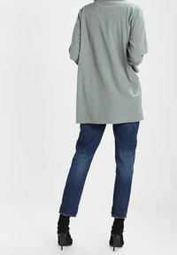 Un cappotto lungo di un grigio chiaro con una vestibilità rilassata, realizzato in un tessuto morbido, abbinato a jeans in denim blu scuro e stivali col tacco neri.