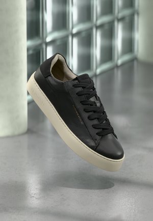 Czarny skórzany sneaker z beżową podeszwą i czarnymi sznurowadłami, unoszący się nad gładką betonową podłogą w pobliżu ściany z bloków szklanych i białej kolumny.