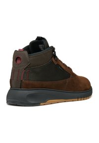 Zapato de caña alta de ante marrón y malla con suela de goma negra, que presenta acentos rojos y un ojal circular cerca del talón para un diseño adicional.