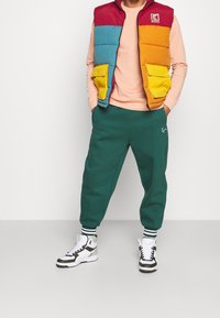 Barevná péřová vesta s červenými, modrými, oranžovými a žlutými částmi, nošená přes růžový top a kombinovaná se zelenými tepláky a bílými teniskami.