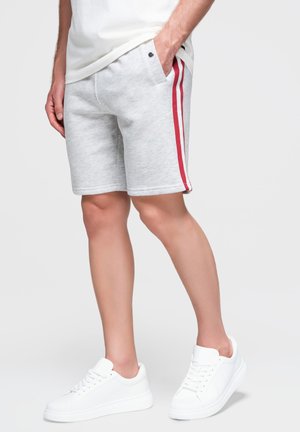 Personne portant un short gris clair avec des bandes rouges sur les côtés, des baskets blanches et une chemise blanche, debout avec une main dans la poche.