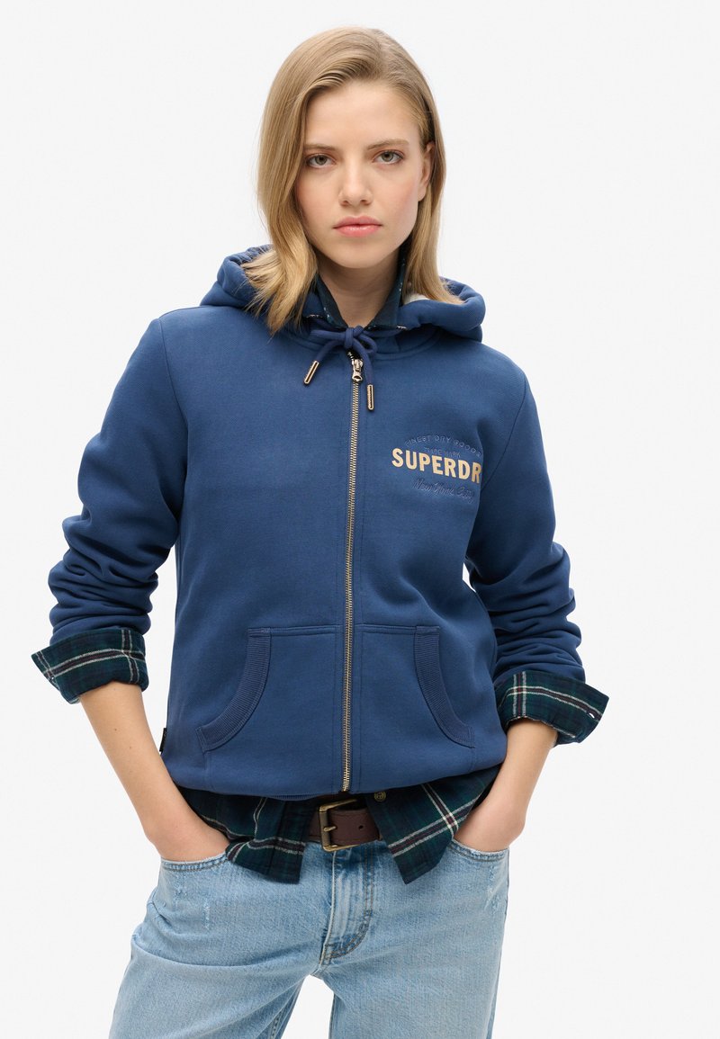 Sudadera con cremallera azul marino y capucha con cordón, que presenta el logo dorado de "SUPERDRY", bolsillos frontales y un cuello de camisa de cuadros asomando.