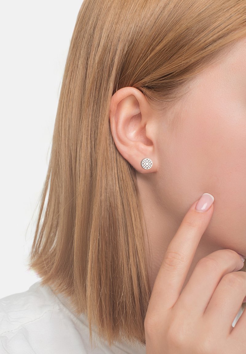 Silver studörhängen med invecklade cirkulära utskärningar, placerade mot en ljus hudton och kort blont hår, som framhäver textur och design.