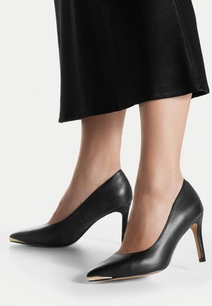 Donna che indossa décolleté nere in pelle con punta a punta abbinate a una gonna nera, in piedi su uno sfondo bianco.