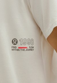 T-shirt de sport blanc avec texture, portant un texte imprimé : "1998 TROUVEZ LE FLUX DANS LE VOYAGE" avec des accents noirs et rouges.