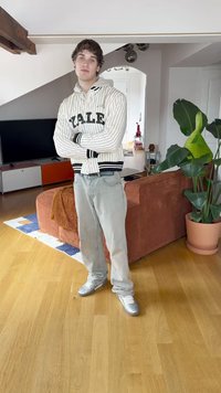 Sudadera a rayas en blanco y negro con el texto "Yale", jeans grises y zapatos plateados metálicos. De pie en una habitación con una planta y un sofá.