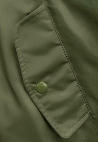 Alyvinės žalios spalvos striukės dalis su užklijuota kišene ir spaudžiamuoju mygtuku, demonstruojanti švelnią audinio tekstūrą ir kontrastingo siuvimo detales.